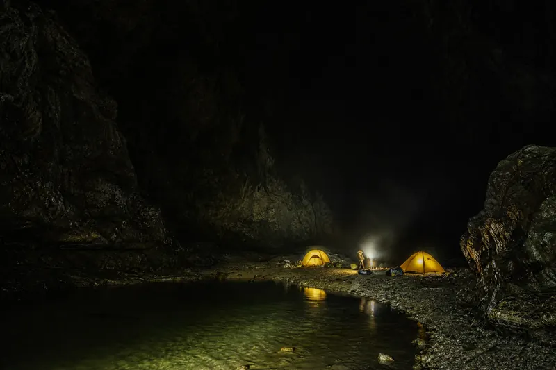 巨大洞厅‍内两顶发光帐篷组成的地下营地，队员在暗河边生火做饭